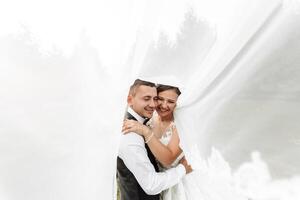 A bride and groom are hugging each other under a white veil. The bride is wearing a white dress and the groom is wearing a black vest. Scene is happy and romantic photo