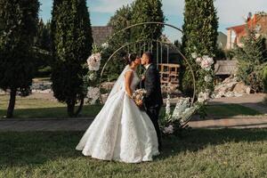 A bride and groom kiss in front of a wedding arch. The bride is wearing a white dress and the groom is wearing a suit. The arch is decorated with flowers and the couple is surrounded by greenery photo
