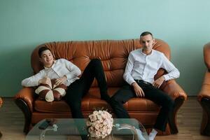 Two men are sitting on a brown couch, one of them is holding a pillow. The couch is surrounded by other furniture, including a coffee table and a chair. Scene is relaxed and comfortable photo
