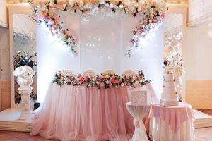 A wedding reception with a pink tablecloth and floral decorations. A cake is on the table and there are chairs around it photo