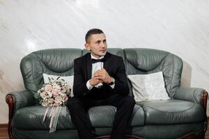 A man in a suit sits on a couch with a bouquet of flowers in his lap. He is wearing a black tie and a white shirt. The couch is green and has a pillow on it photo