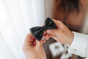 A man is holding a black bow tie in his hand. The bow tie is shiny and looks well-made. The man seems to be getting ready for a formal event, such as a wedding or a business meeting photo