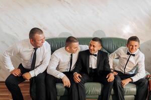 Four men are sitting on a couch, all wearing black suits and ties. They are smiling and seem to be enjoying each other's company. Scene is lighthearted and friendly photo