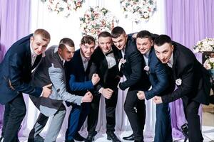 A group of men in suits are posing for a picture. Scene is happy and celebratory, as they are all dressed up and posing for a group photo