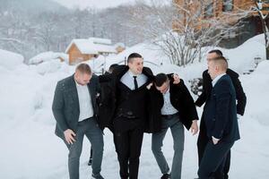 A group of men in suits are posing for a picture in the snow. They are all smiling and seem to be enjoying themselves. Scene is lighthearted and fun, as the men are embracing each other photo