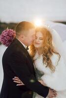 A bride and groom are embracing each other in the snow, with the bride wearing a white fur coat and the groom holding a bouquet of pink flowers photo