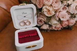 A white box with a red velvet lining and a heart on it. Inside the box are two rings. A bouquet of pink flowers sits next to the box photo