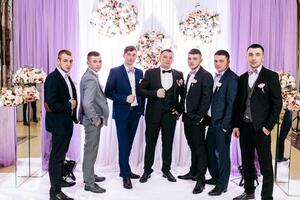 A group of men are posing for a photo in front of a purple backdrop. They are all dressed in suits and ties, and one of them is holding a camera. Scene is formal and professional