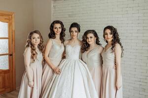 A group of five women are posing for a picture in front of a white wall. They are all wearing dresses and are smiling for the camera. Scene is happy and celebratory photo