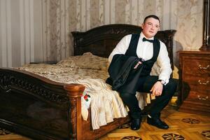 A man in a suit and tie sits on a bed. He is holding a briefcase and he is getting ready for a formal event photo