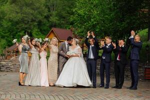 A group of people are posing for a picture, including a bride and groom. The bride is wearing a white dress and the groom is wearing a suit. The group is standing in front of a building photo