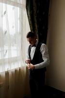 A man in a suit is standing in front of a window, adjusting his tie. Concept of formality and attention to detail, as the man takes the time to ensure his appearance is polished and professional photo