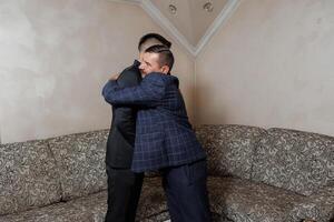 Two men hug each other in a living room. One of them is wearing a suit. Scene is warm and friendly photo