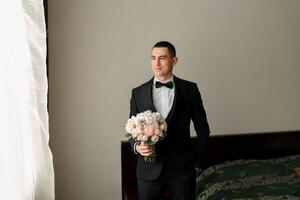 A man in a suit and bow tie stands in front of a bed with a bouquet of flowers in his hand photo