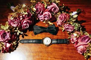 A watch and a black bow tie are displayed on a table with a bunch of purple flowers. The watch is a gold and black one, and the flowers are arranged in a circular pattern photo