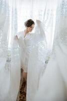 A woman in a white robe stands in front of a curtain, with a lace veil on her head photo