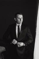 A man in a suit is standing in front of a window, adjusting his tie. Concept of formality and professionalism, as the man is dressed in a suit and tie photo