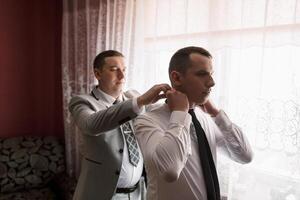 Two men are getting dressed up, one of them is wearing a tie. The man in the suit is helping the other man tie his tie photo