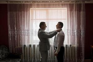 Two men are getting dressed in front of a window. One man is helping the other tie his tie photo