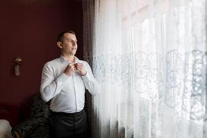 A man in a white shirt is getting dressed in front of a window. The curtains are white and have a floral pattern. The man is smiling as he gets dressed, giving the impression that he is happy photo
