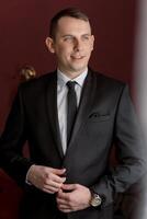 A man in a suit and tie is standing in front of a red wall. He is wearing a watch and has a smile on his face photo