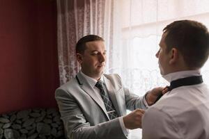 Two men are getting dressed up, one of them is adjusting the tie of the other. The man adjusting the tie is wearing a gray suit and tie, while the other man is wearing a white shirt photo