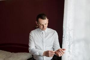 A man in a white shirt is getting ready for a formal event. He is adjusting his tie and checking his reflection in the window. Scene is one of preparation and anticipation for the upcoming event photo