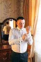 A man in a white shirt is getting dressed in front of a mirror. He is adjusting his cufflinks and looking at himself in the mirror photo