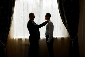 Two men are getting dressed up in front of a window. One man is helping the other man tie his tie photo