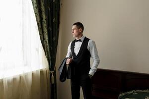 A man in a black vest and a black bow tie is standing in front of a window. He is holding a black briefcase in his hand. Concept of formality and professionalism photo