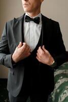 A man in a black suit and white shirt is getting ready to wear a black bow tie. He is adjusting his suit jacket and tie, and his hands are visible. Concept of formality and elegance photo