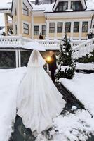 A bride and groom are standing in the snow, with the bride holding an umbrella. The scene is set in front of a yellow house, and the couple appears to be getting ready for their wedding photo