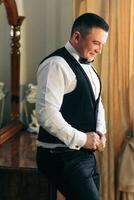 A man in a black vest and white shirt is standing in front of a mirror. He is adjusting his tie and he is in a good mood photo