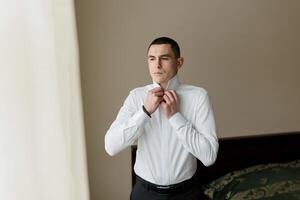 A man in a white shirt is getting dressed and adjusting his collar. Concept of formality and attention to detail, as the man takes care to ensure his appearance is neat and polished. The setting photo
