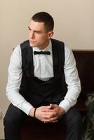 A man in a black vest and white shirt is sitting on a bed. He is wearing a black bow tie and has his hands clasped in his lap. Concept of formality and elegance photo