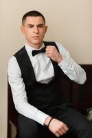A man in a black vest and white shirt is sitting on a bed. He is adjusting his tie and looking at the camera. Concept of formality and elegance, as the man is dressed in a suit and tie photo