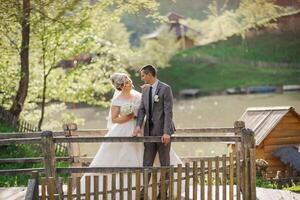 A bride and groom are standing on a wooden fence overlooking a lake. The bride is holding a bouquet and the groom is wearing a tie. The scene is peaceful and romantic photo
