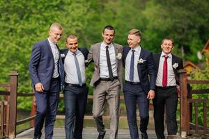 A group of five men dressed in suits and ties are posing for a photo on a bridge. Scene is lighthearted and fun, as the men are smiling and enjoying each other's company