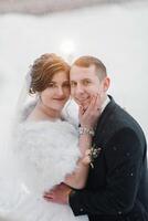 A bride and groom are posing for a picture in the snow. The bride is wearing a white fur coat and the groom is wearing a black suit. They are both smiling and seem happy to be together photo