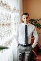 A man in a white shirt and gray pants stands in front of a window with a plant behind him. He is wearing a black tie and has his hands in his pockets. Concept of professionalism and formality photo