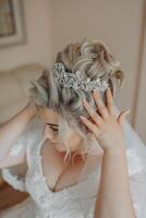 A woman is getting ready for a wedding and is putting on a tiara. The tiara is made of gold and has a lot of diamonds. The woman is wearing a white dress and is standing in front of a couch photo