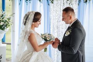 A bride and groom are getting married in front of a blue curtain. The bride is wearing a veil and the groom is wearing a suit photo