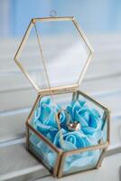 A gold and blue box with a blue rose and two gold rings inside. The box is placed on a table photo