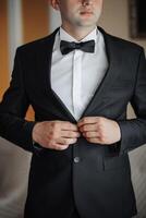 A man in a black suit and white shirt is getting ready to tie his bow tie. Concept of formality and elegance, as the man is dressed in a tuxedo and preparing for a special occasion photo