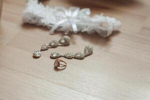 A wedding ring and a pair of earrings are displayed on a wooden table. The earrings are made of diamonds and are placed on top of the ring photo