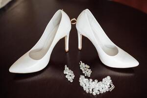 A pair of white shoes with a ring on the back and a pair of pearl earrings. The shoes are placed on a table with a black background photo