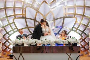 A couple is kissing in front of a table with drinks and flowers. The table is set for a wedding reception photo