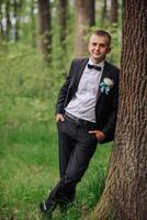 A man in a suit and tie stands next to a tree. He is wearing a blue and white boutonniere photo