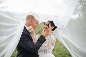 A bride and groom are standing under a white veil. The bride is wearing a white dress and the groom is wearing a black suit. They are both smiling and looking at the camera photo