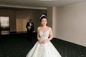 A bride and groom are standing in a room with a large white dress on the bride. The bride is wearing a tiara and the groom is wearing a tuxedo photo